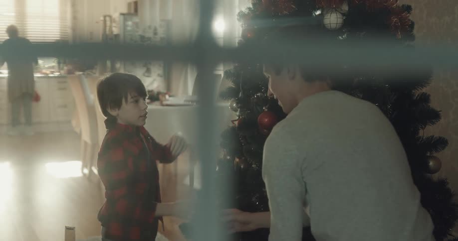 Two people unbox ornaments and add them to christmas tree while woman works in kitchen