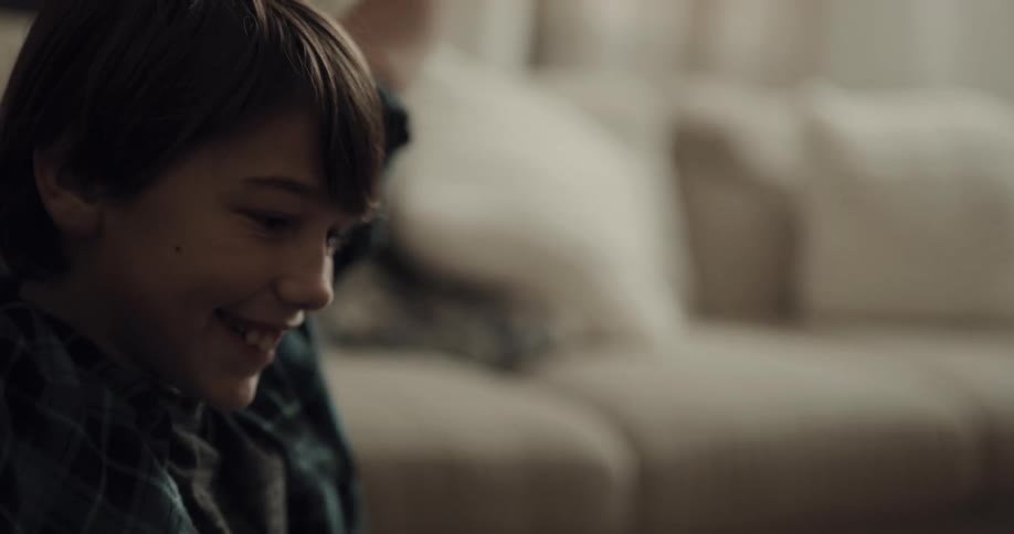 A young boy smiles and raises his arms up as he sits on the floor in front of the couch in his living room