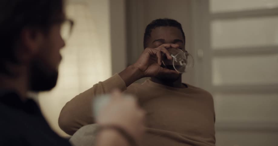 Two men sit in a house and talk while one drinks a glass of wine
