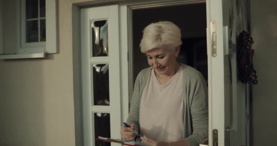 A delivery man carrying packages knocks on a door and a woman answers and signs for the packages as a young boy brings the man a gift