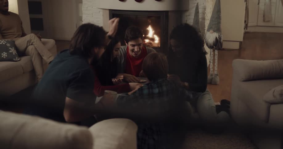 A group of friends laugh as they sit around a coffee table and play cards in front of a fireplace