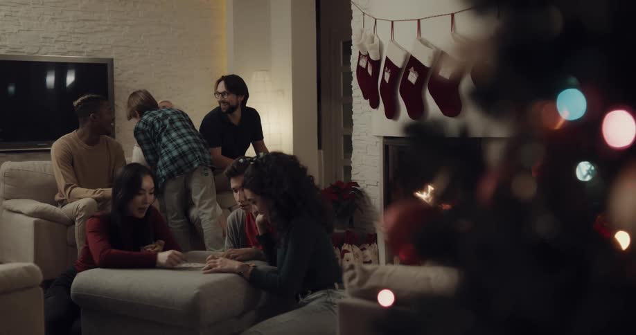 A young boy takes a gift from under a christmas tree and runs and gives it to his grandmother who sits with two others as three other people play a game