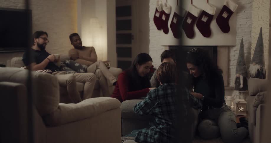 A group of friends sit around in a living room by the fireplace and hung christmas stockings