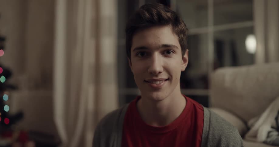 Teenage boy looks and smiles at the camera next to the christmas tree in the living room at home