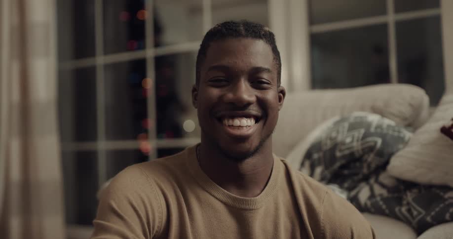 Man with a brown sweater sits next to the couch and faces the camera with a broad smile and eye contact