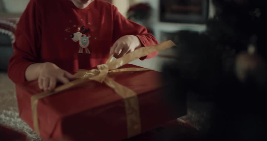 Male child sitting next to the christmas tree holds a wrapped christmas present up to his ear while shaking it and then places it on his lap while untying the ribbon to unwrap the gift