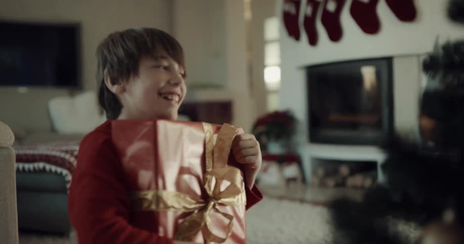 A young boy takes a present from under a christmas tree and shakes it and starts to open it