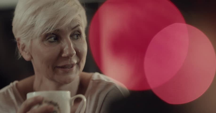 A woman with short white hair drink from a glass coffee mug while smiling and conversing