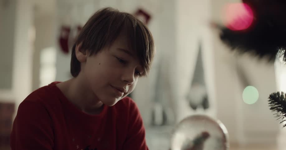 A boy sitting in a room next to a christmas tree holding a snow globe