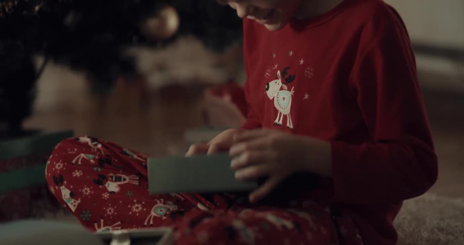 A boy retrieves and opens a gift from beneath a christmas tree before rising to hug his grandmother