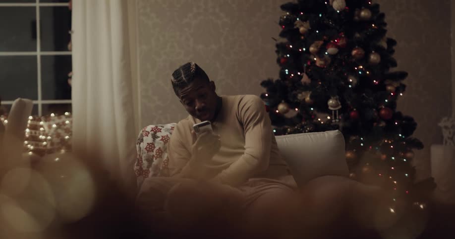 Man sits in chair near christmas tree laughing while video calling via mobile phone