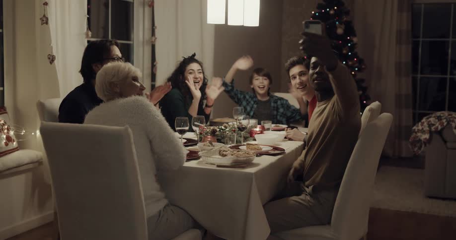A family sits at a table at home and a man pulls his cell phone out and they all wave for a selfie