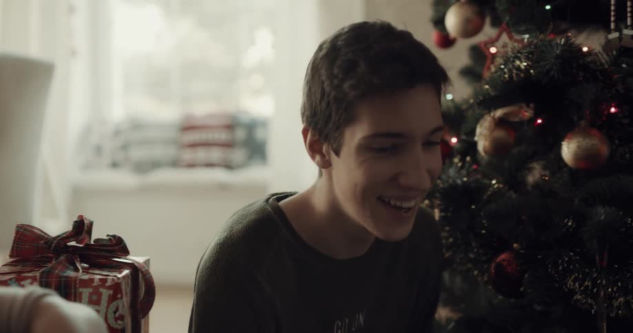 A boy takes a gift from under the christmas tree and opens it as his brother and grandmother watch