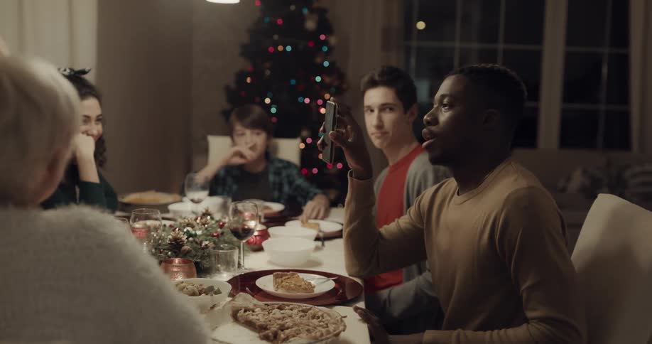 A group of people sit around a family dinner table and video call someone on a cell phone