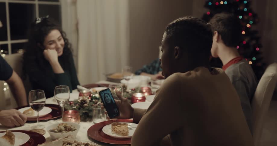 A group of people sit at the dinner table as a young man video chats with a woman on his cell phone