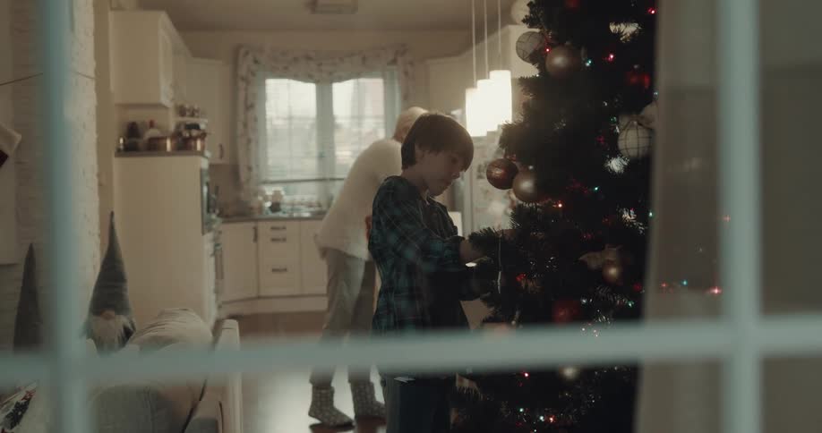Boy decorated christmas tree while grandmother carries plate from kitchen