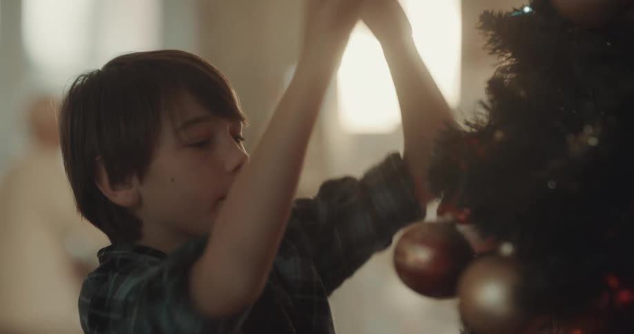 Boy decorates christmas tree as people move around in the room behind him
