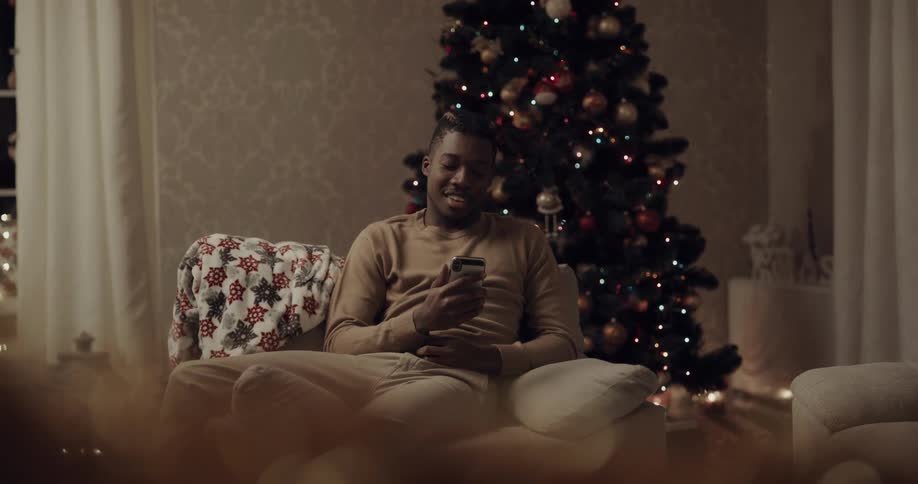 Man with a brown sweater and pants sits in an armchair next to the christmas tree and makes a video call on his smartphone in the living room at home