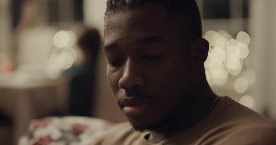 A black male sitting indoors casts his gaze downwards while talking to someone