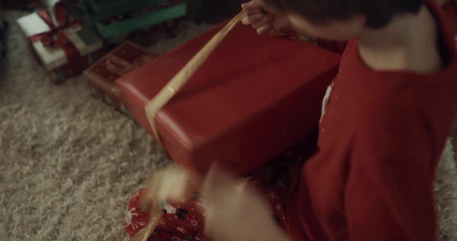 A boy sitting on the floor retrieves a present from beneath a christmas tree and unwraps it