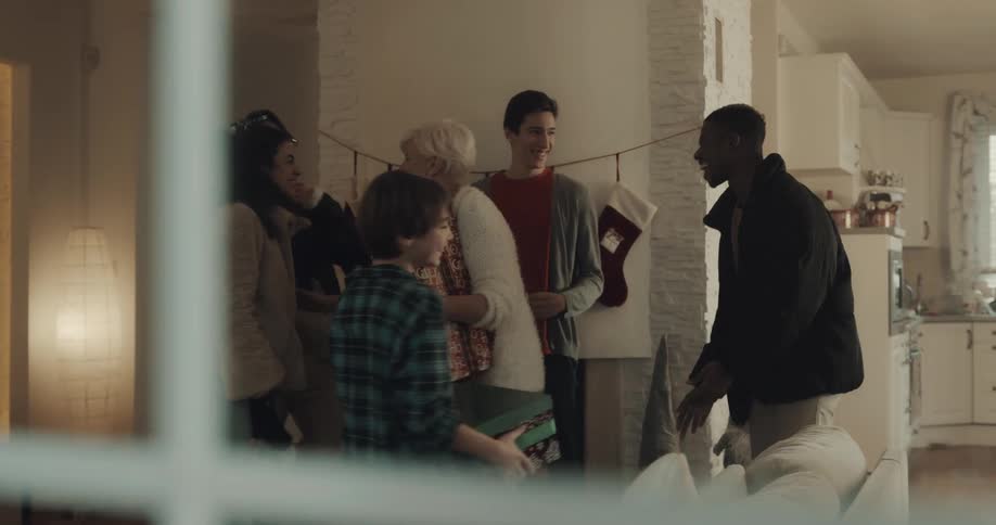 A woman holding gifts hug a man and woman as they come in to her home and others in the house high five one another