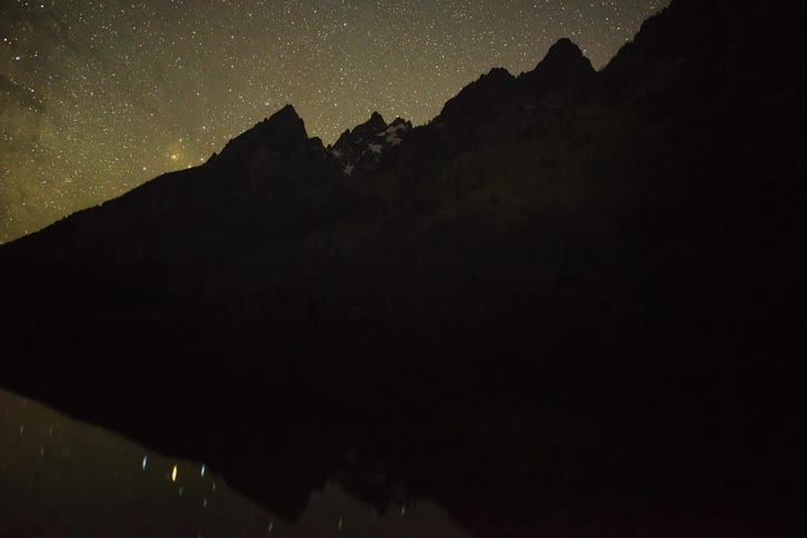 A time lapse of a a river and mountains during the day and then at night as stars cover the sky