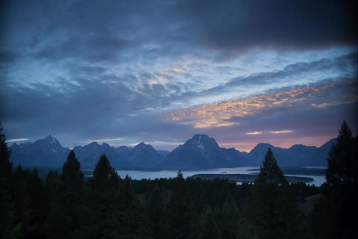Clouds pass by at a sped up pace as the sun sets over a mountain backdrop surrounded by lakes and a forest