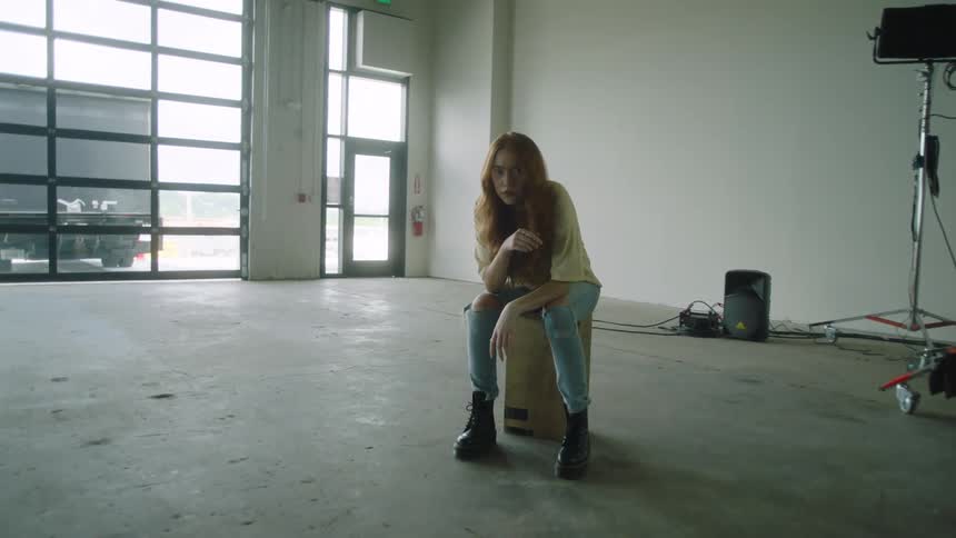 A woman sits on a box in an empty work space with large windows