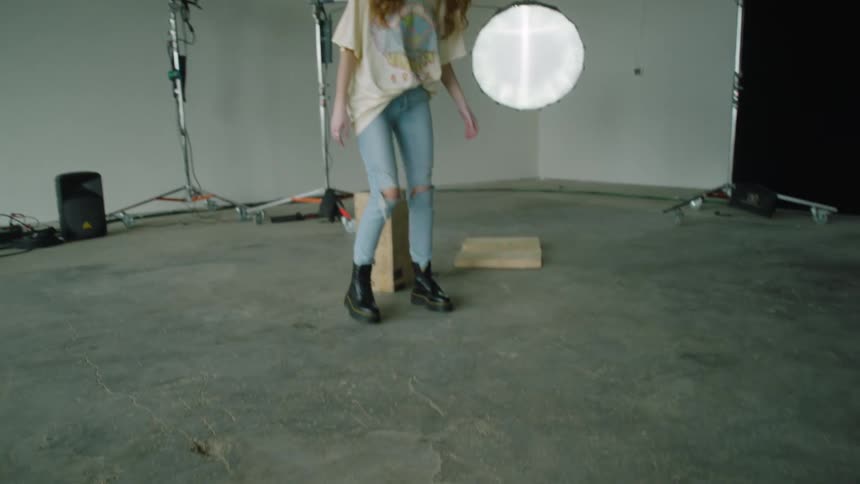 A woman sitting on a box in a room with a wall a speaker and wires in the background