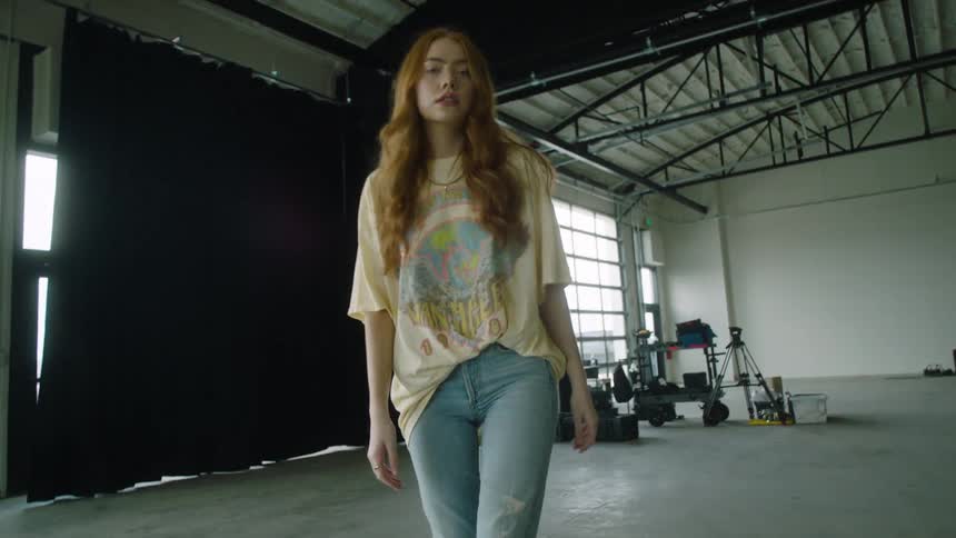 A young woman walks backwards through a photography studio while looking into the camera before turning her back