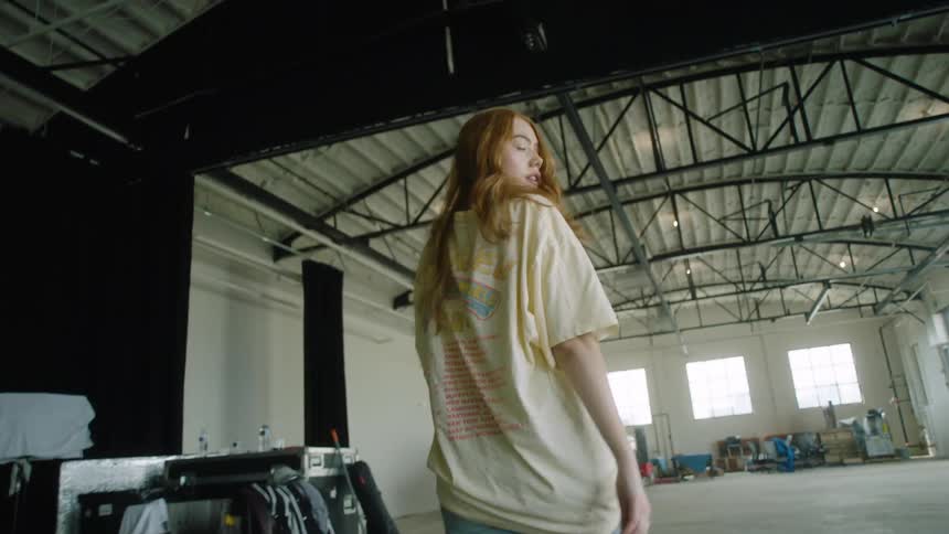 A young woman walks across a warehouse while looking behind her from right to left