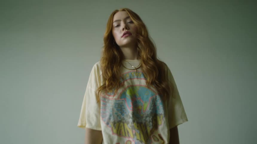 A female teenager with red hair wearing a t shirt and jeans stands in front of a grey wall