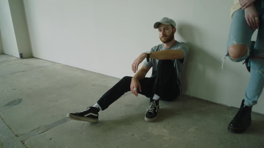 Man wearing a hat sits on the floor and leans against a wall while another person stands nearby