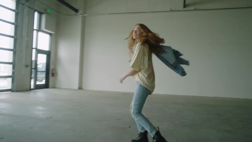 Model in jeans a t shirt a necklace boots and holding a jean jacket walks backwards and spins then throws the jacket smiling in the background there are windows a door and an exit sign a fire extinguisher a blank wall and black curtains a truck and car