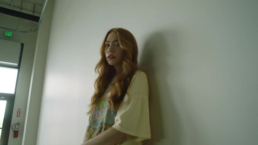 A woman standing and staring next to a wall with a door and exit sign in the background