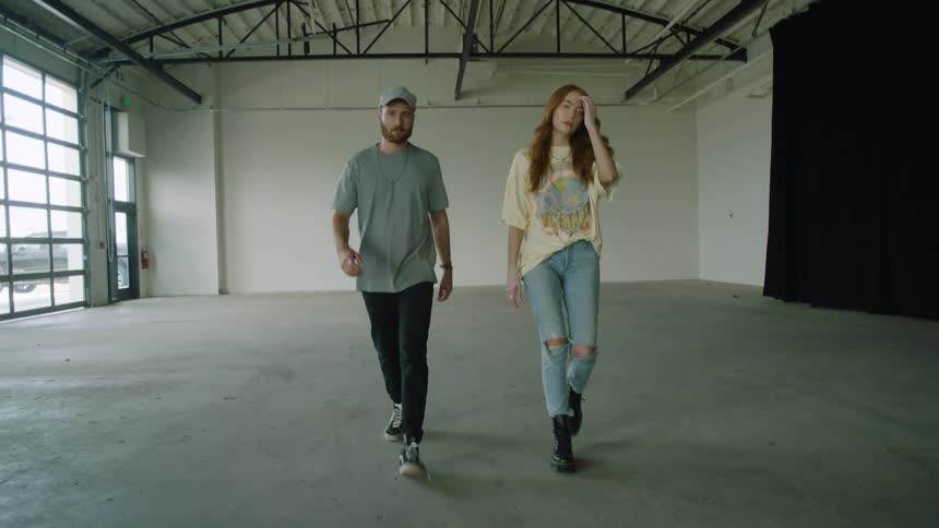 Man and woman walk across the floor of a large empty room as the woman pushes her hair back with her hand