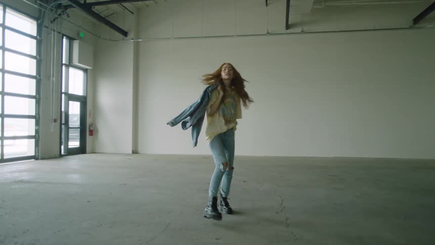 A young woman walks across a large empty room while peaking back at the camera as it follows her