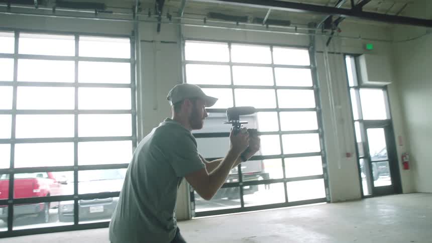 A man is filming a woman walking around in an empty garage with vehicles parked outside and natural light coming inside the garage