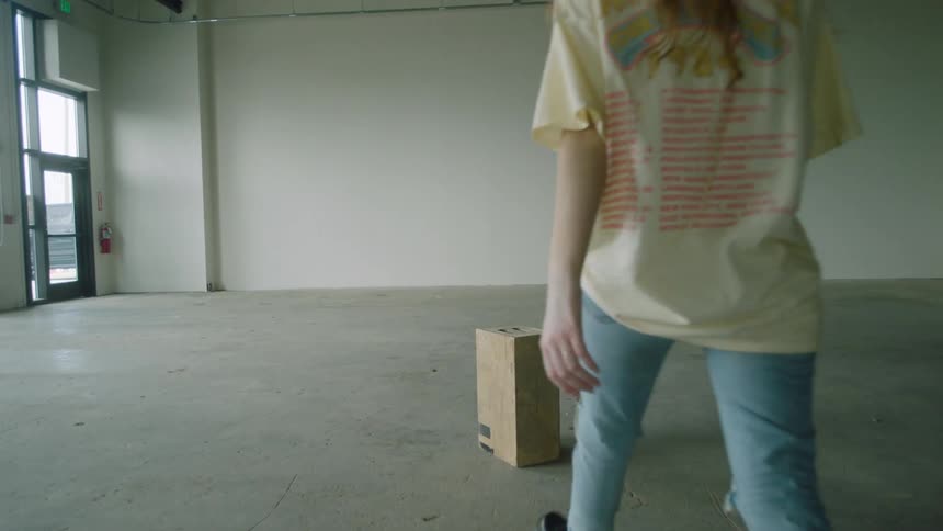 A girl walks to a box in the middle of a room and sits on it