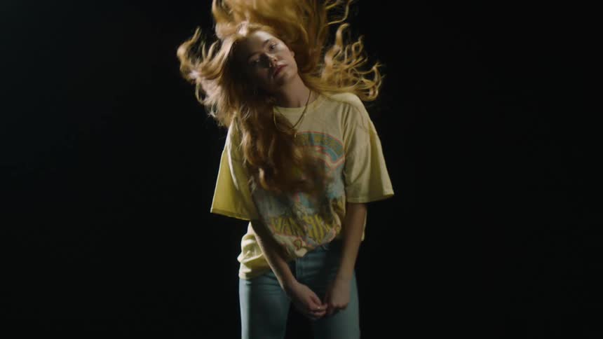 Blonde young woman whips her long hair from side to side while looking at the camera in a dark room