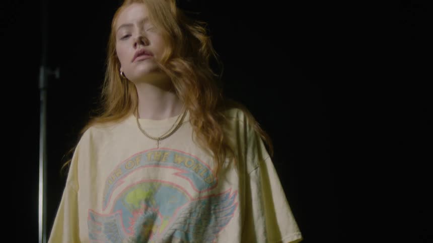 A young woman poses in a dark studio wearing an oversized yellow t shirt