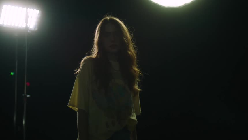 Woman with long hair stands in a dark room as light shines in the window behind her