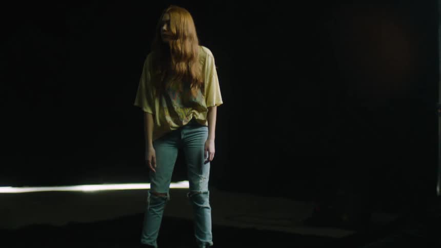 Model poses standing with hair covering most of face in dark room with few lights in boots jeans and a t shirt