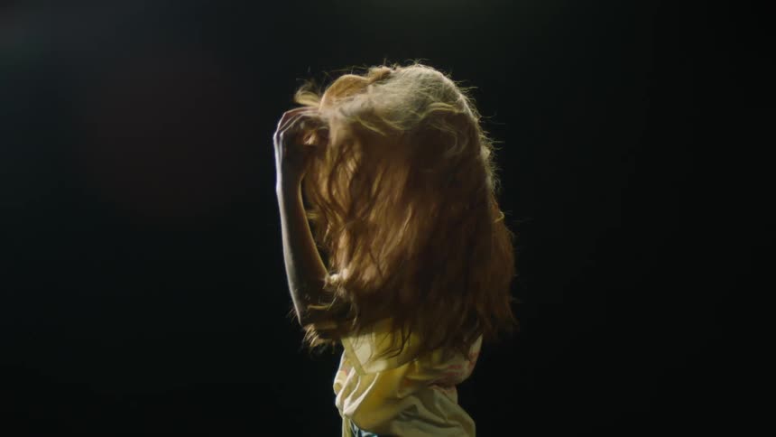 A young woman positioned to her side flips her hair upwards and then stares into the camera
