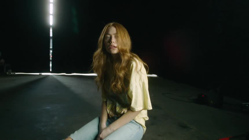 A young woman poses on a concrete block in a dark studio with her head cocked to the side