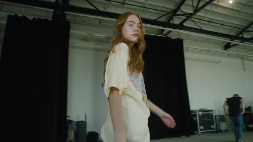 Model is sitting on a wooden block gets up then walks towards other people working a truck and equipment some of which is in cases in a warehouse type building with black curtains a metal roof and rafters past exit signs glass doors and windows