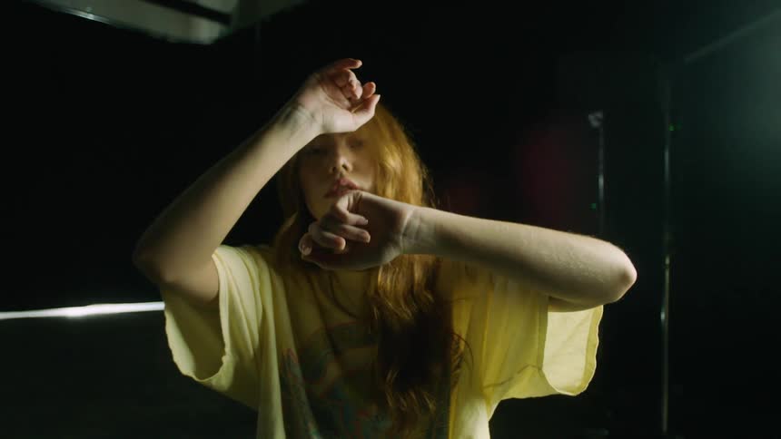 A teenage girl sits on a stool in front of a bright light and moves her arms around her face