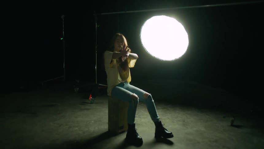 Woman sitting on box in an empty studio moves rhythmically and gestures at camera