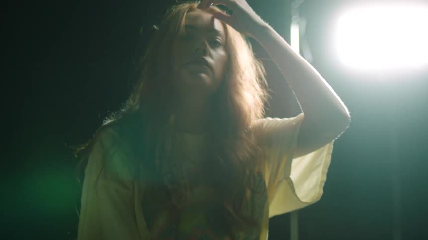 A young woman sitting before a spotlight in a dark studio runs her hand through her hair as she looks into the camera