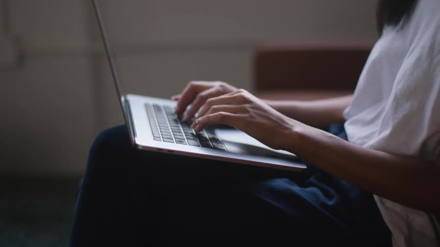 A young woman types on a laptop while sitting on a brown couch